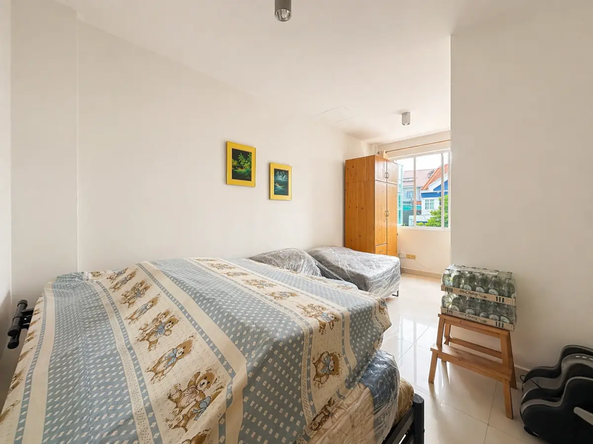 Bright bedroom with two single beds, a tall wooden wardrobe by the window, and a crate of canned drinks on a small stand.