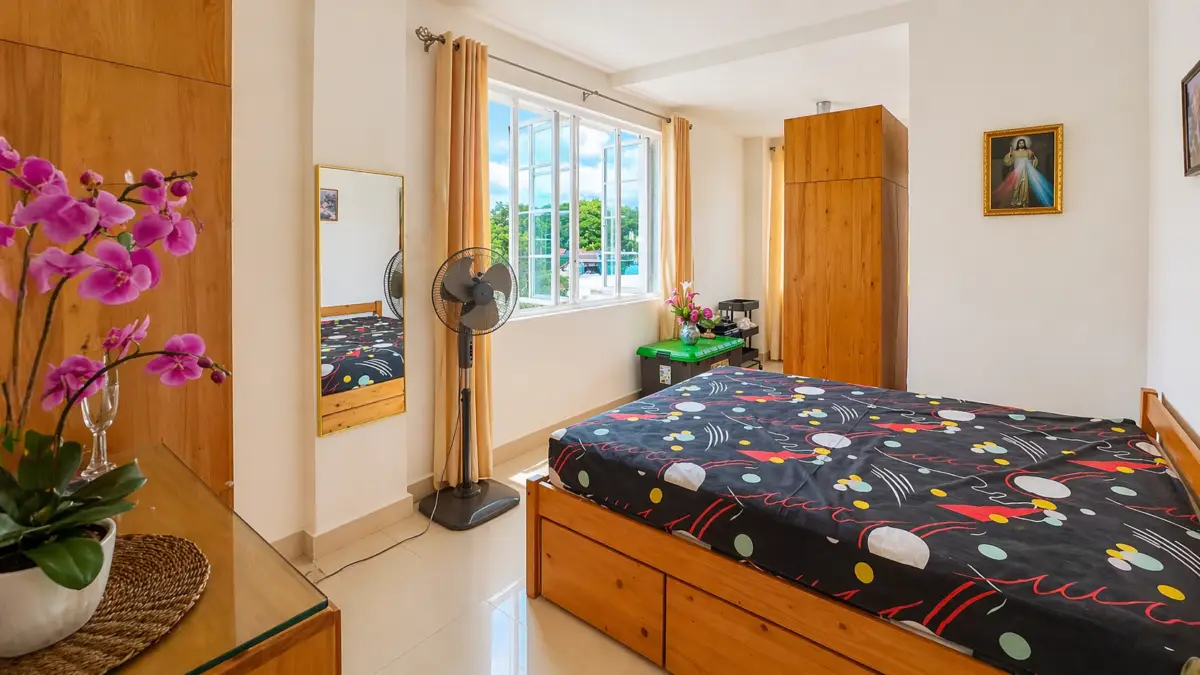Bright bedroom with a wooden bed and matching wardrobes, a colorful patterned bedspread, and a large window with beige curtains letting in daylight.