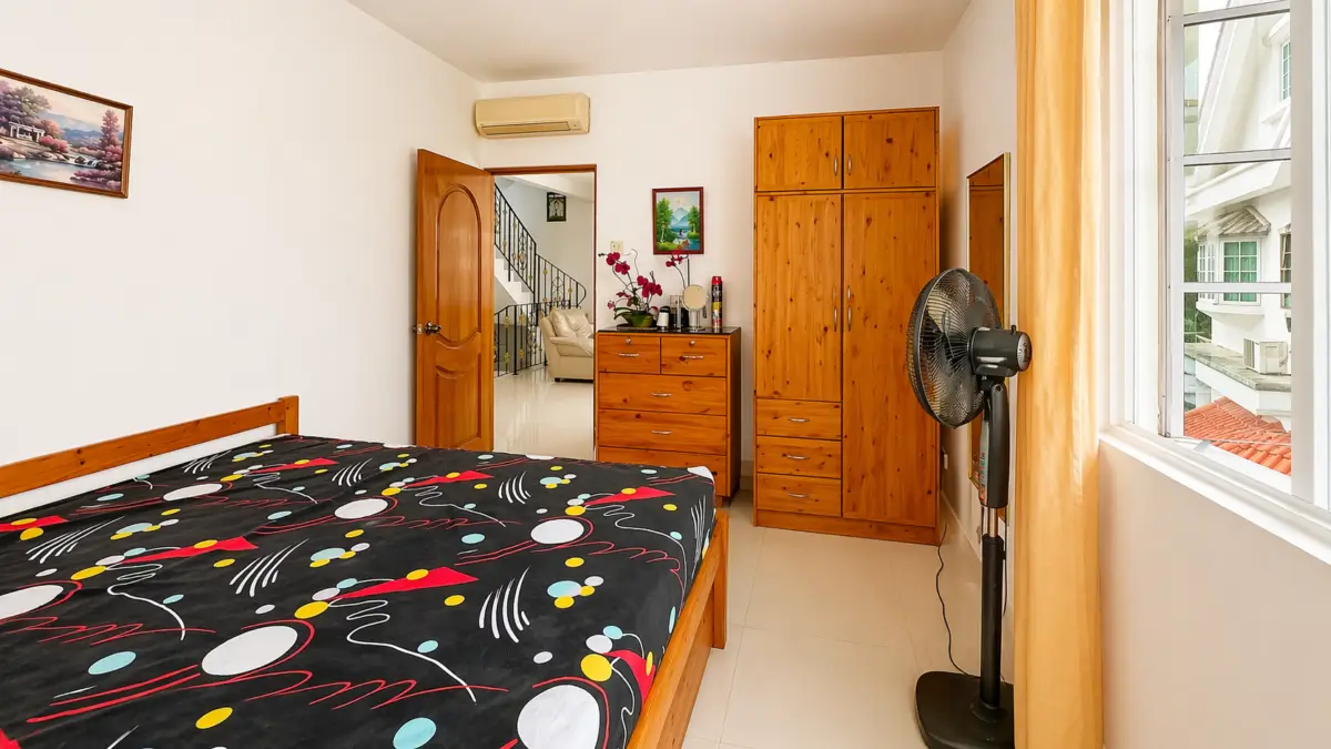 Bedroom with a wooden bed, colorful patterned duvet, dresser, wardrobe, and a standing fan by a large window with yellow curtains.