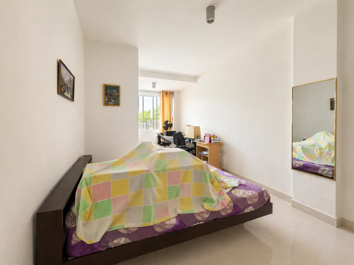 A small bedroom with a low wooden bed, colorful patchwork blanket, and a large wall mirror on the right.