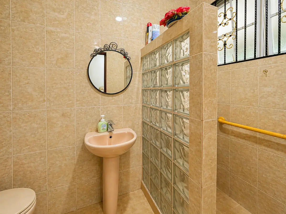 Beige-tiled bathroom with a pink pedestal sink and round decorative mirror above it, soap dispenser on the sink, and a glass block divider to the right.
