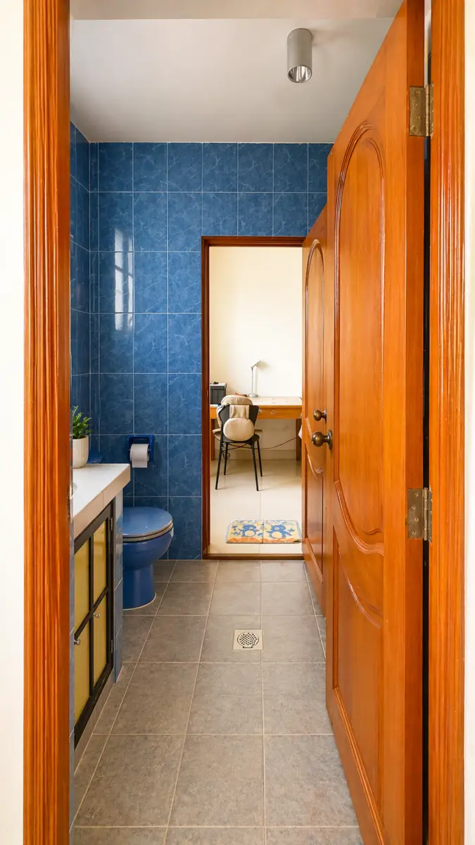 Blue-tiled bathroom with a blue toilet and sink, open wooden door leads to a study with a desk and chair.