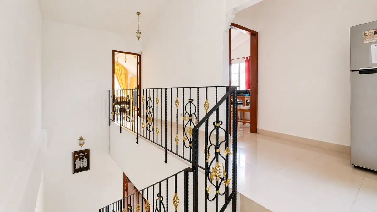 Bright two-level hallway with white walls, black wrought-iron railing with gold accents, and open doorways to rooms; a stainless refrigerator is visible on the right.