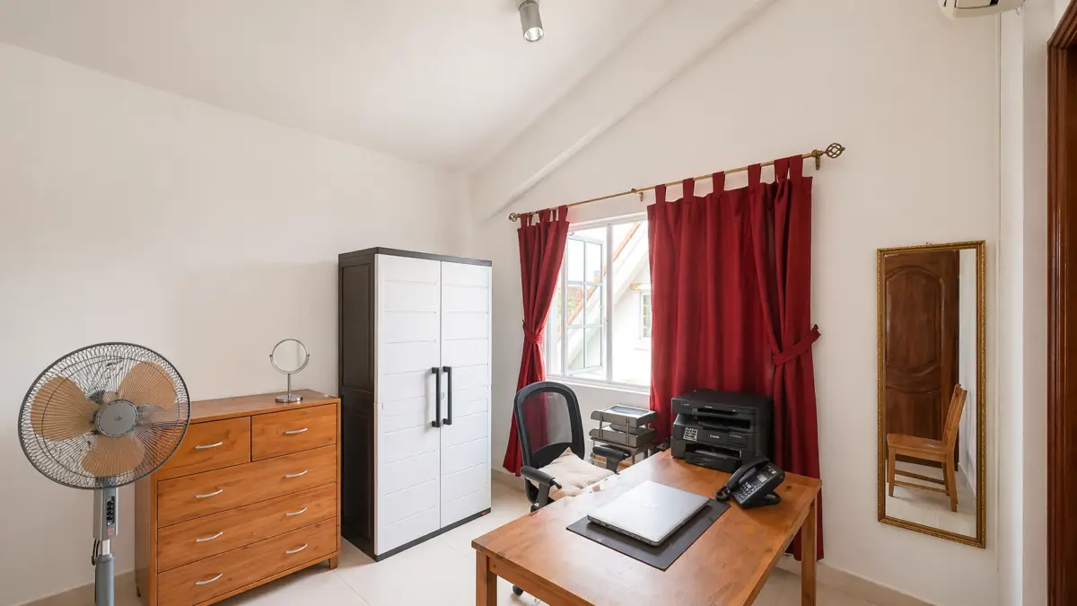 A small home office with a wooden desk, laptop, phone, and printer near red curtains by a window.