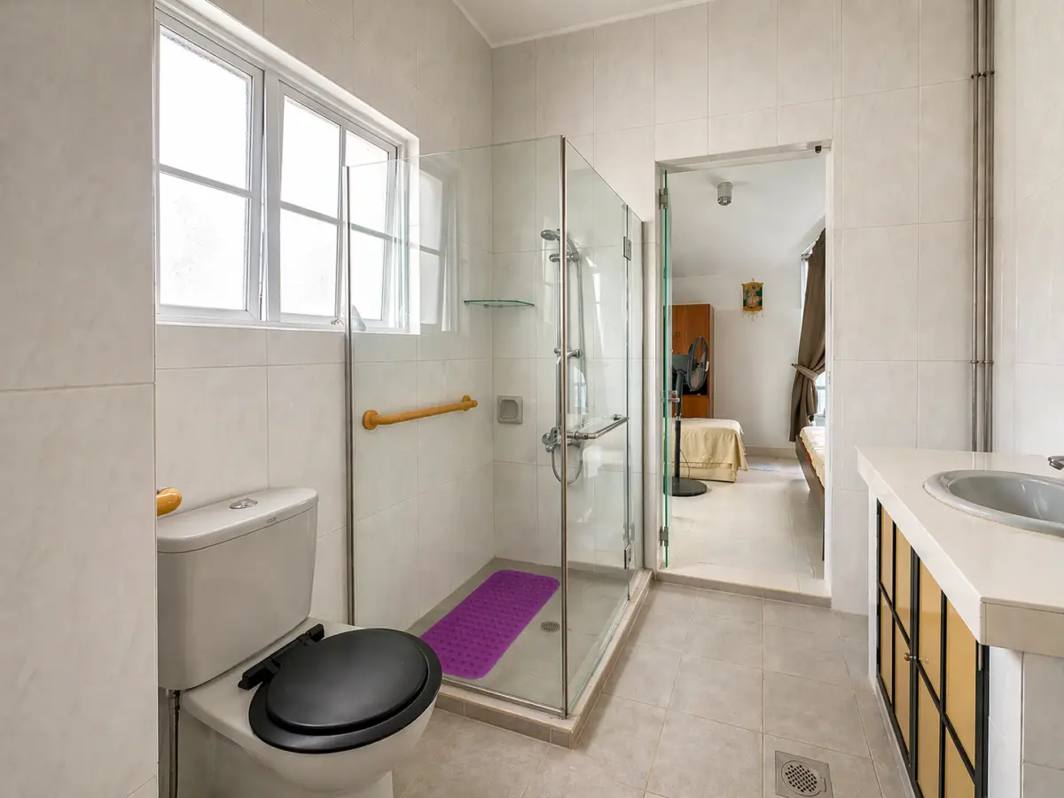 Bright tiled bathroom with a glass shower enclosure, toilet with a black seat, and a purple bath mat; doorway opens to a bedroom.