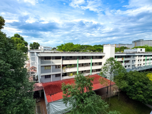 Aerial view of a white multi-story campus building with green vertical accents, red-roofed pavilion, and surrounding trees under a blue sky.