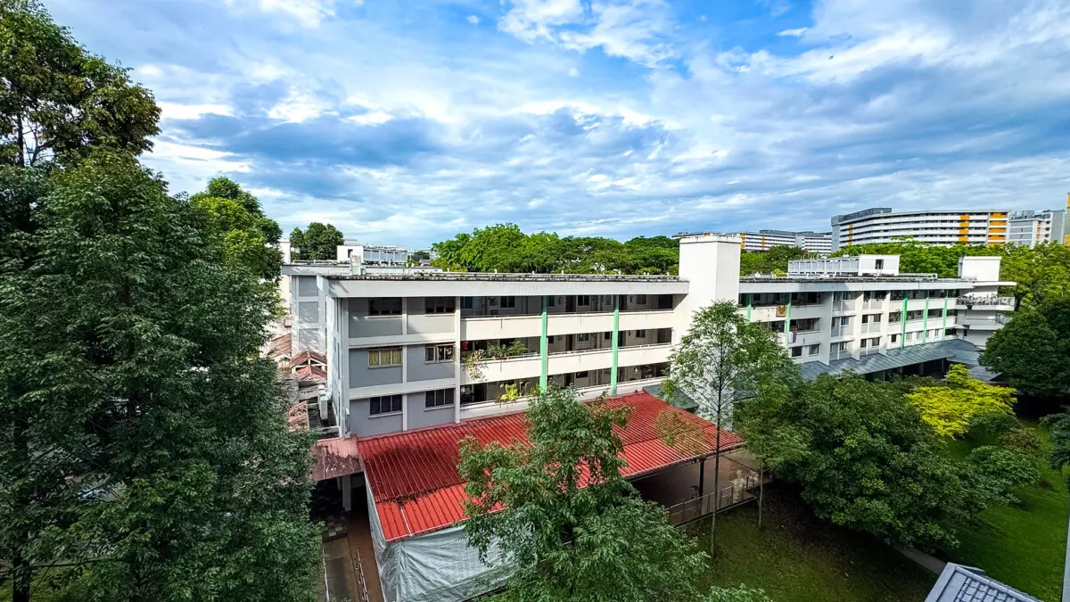 Aerial view of a white multi-story campus building with green vertical accents, red-roofed pavilion, and surrounding trees under a blue sky.