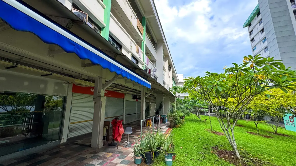 Building exterior with a blue awning over a walkway, closed shop shutters, and a small garden with potted plants and trees.