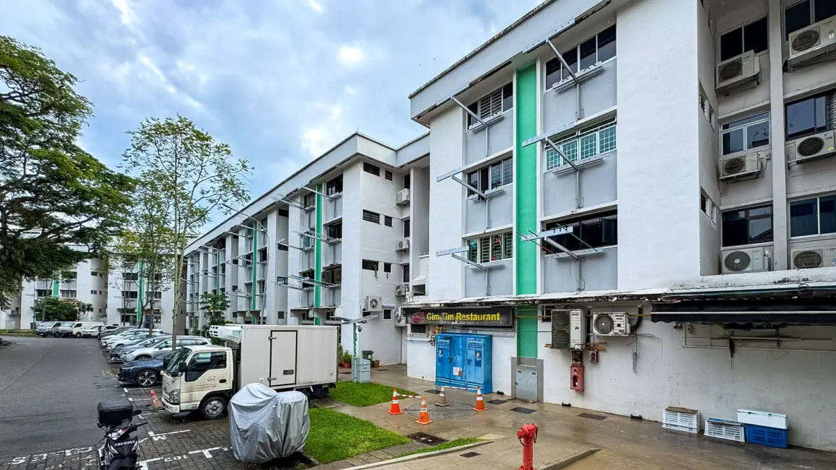White multi-story apartment building with a green vertical accent, ground-floor restaurant storefront, and cars parked along a tree-lined street.