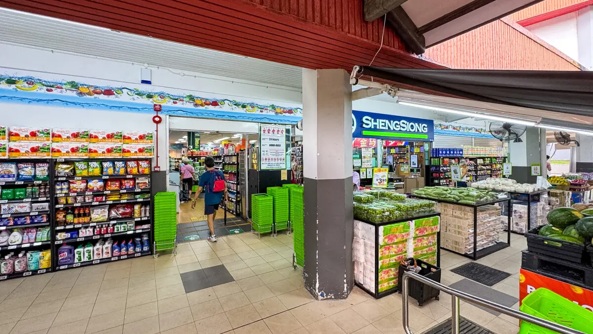 Inside a brightly lit supermarket with shelves of products on the left and fresh produce on the right near a Sheng Siong sign.