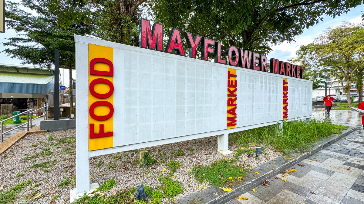 Outdoor sign for Mayflower Market with bold red letters on top and vertical yellow panels labeled FOOD and MARKET; trees and a walkway in the background.