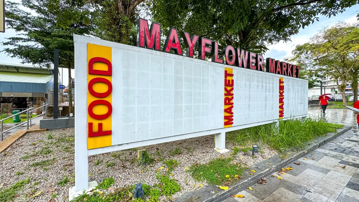 Outdoor sign for Mayflower Market with bold red letters on top and vertical yellow panels labeled FOOD and MARKET; trees and a walkway in the background.