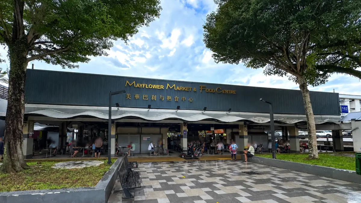 Exterior view of Mayflower Market & Food Centre with outdoor seating, shoppers, and trees along the front.