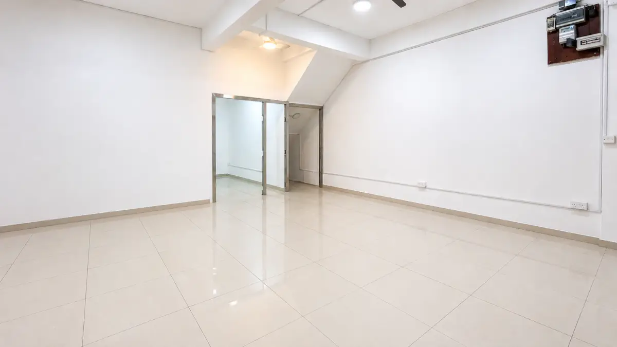 Empty, bright room with glossy beige tiles, white walls, and a glass doorway leading to another area; exposed ceiling beams and a wall-mounted electrical panel on the right.