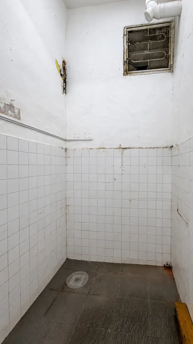 Small, grimy shower stall with white tiled walls, a dark floor, and a round drain cover on the floor.