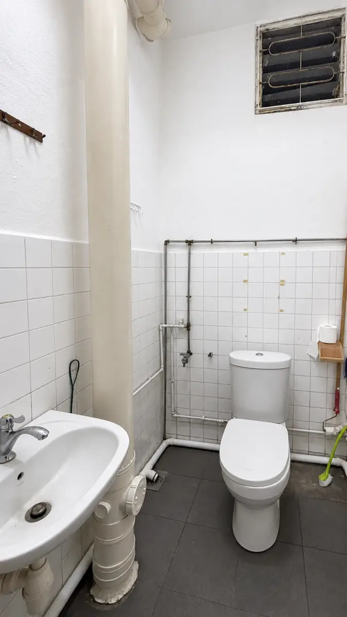 Small utilitarian bathroom showing a white toilet and sink with exposed pipes and a tall vertical vent pipe, white tiles, and a small wall vent near the ceiling.