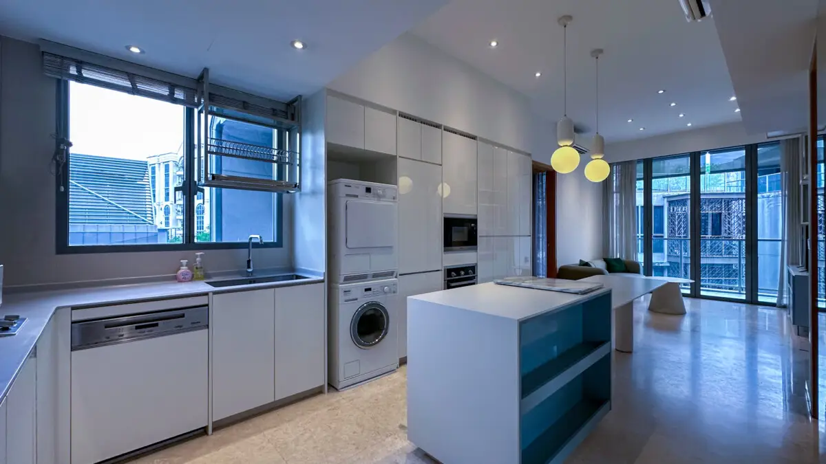 Modern white kitchen with an island, built-in appliances, and a stacked washer and dryer next to tall cabinets in an open-plan space.