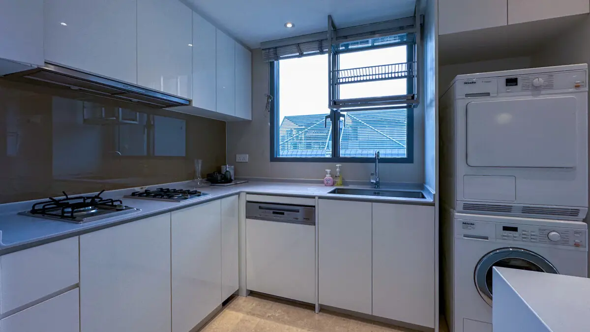 Modern white kitchen with a gas cooktop, built‑in dishwasher, and a stainless steel sink under a large window; stacked washer and dryer on the right.