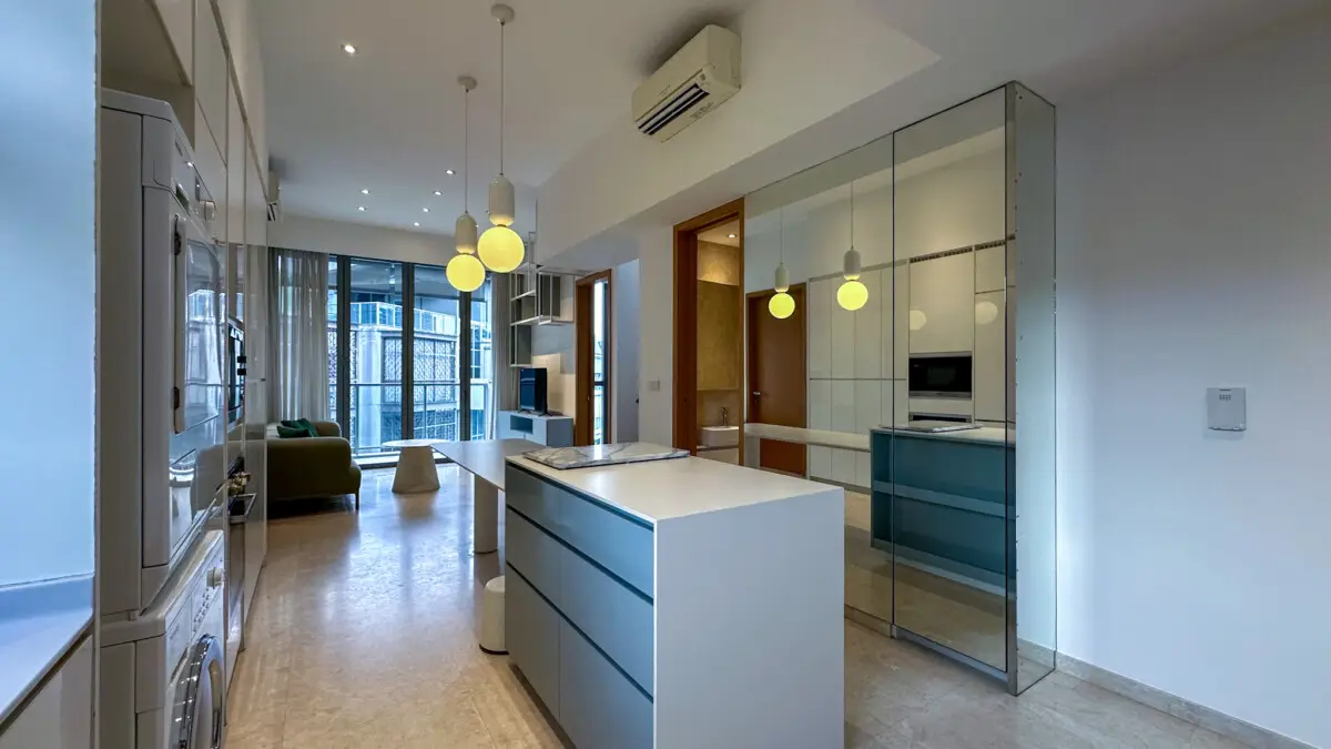 Open-concept kitchen with a white island, blue-drawer cabinets, and three hanging pendant lights leading to a living area with floor-to-ceiling windows.