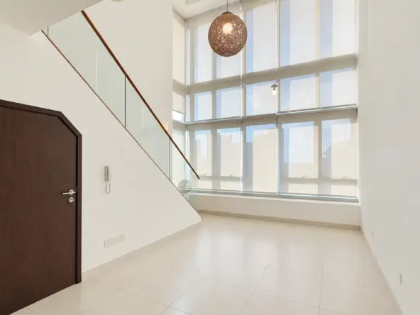 Bright modern interior with a dark wood door on the left, glass-railed staircase, and large windows offering a city view.