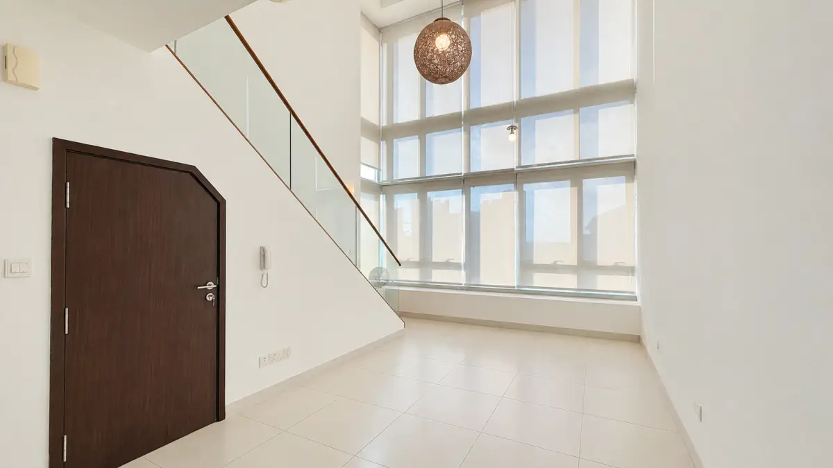Bright modern interior with a dark wood door on the left, glass-railed staircase, and large windows offering a city view.