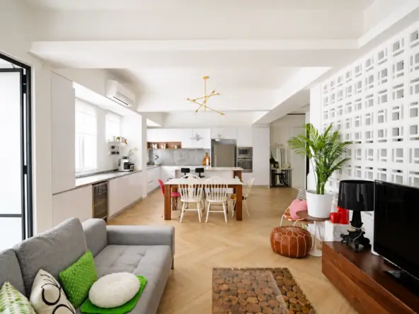 Open-plan living area with a gray sofa, green cushions, and a wooden coffee table facing a TV; bright kitchen in the background and a lattice wall on the right.