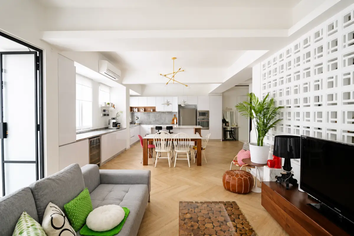 Open-plan living area with a gray sofa, green cushions, and a wooden coffee table facing a TV; bright kitchen in the background and a lattice wall on the right.