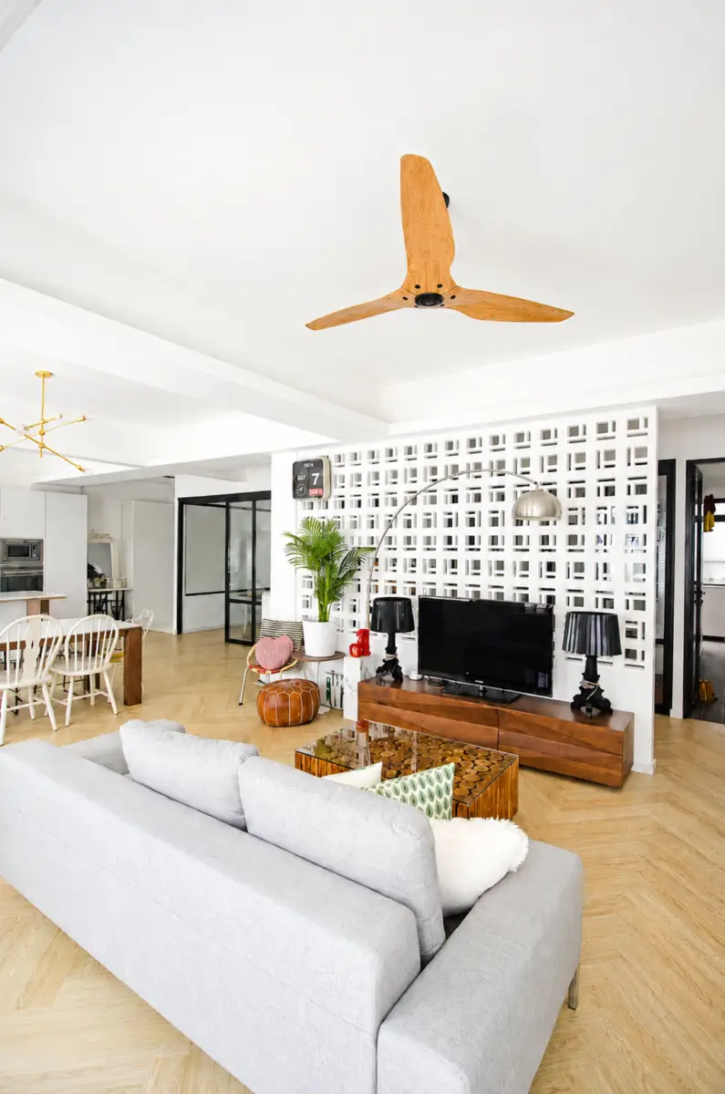 Open-plan living area with a gray sofa facing a wooden TV console, lattice wall, and a large wooden ceiling fan overhead; a dining area and plants visible in the background.
