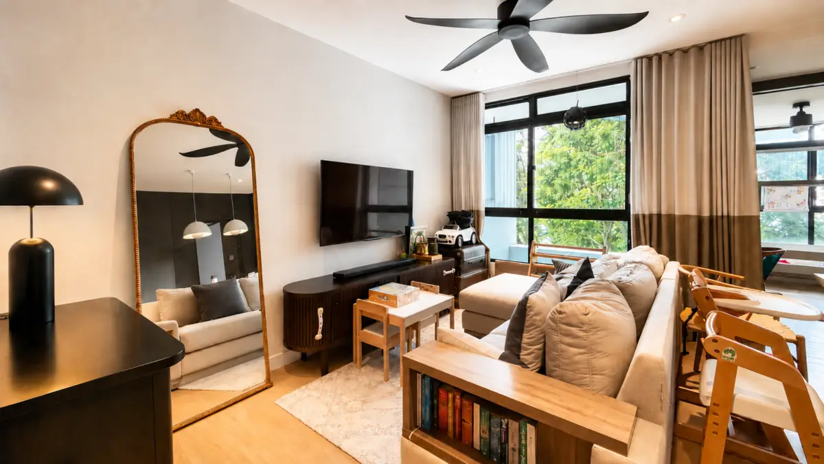 Bright living room with a beige sectional, wooden tables, and a wall-mounted TV; large windows with brown curtains bring in natural light.