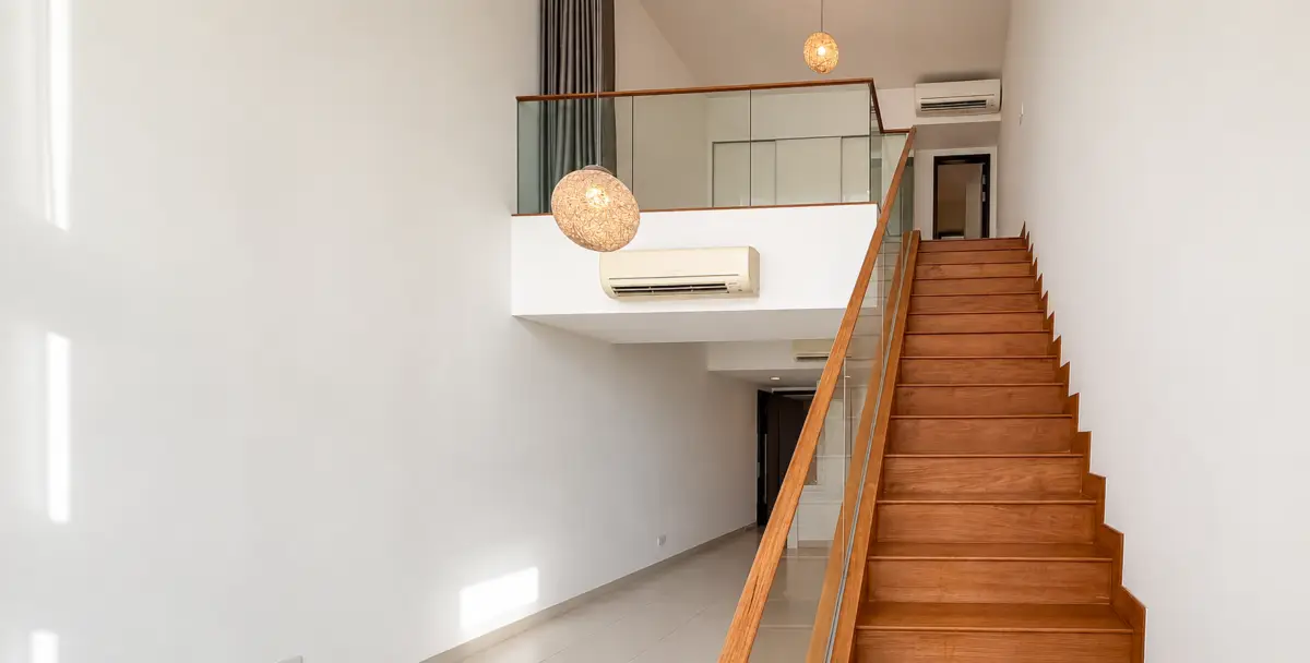 Bright two-story lobby with wooden staircase, glass railing, white walls, and hanging wicker pendant lights at the top balcony area.