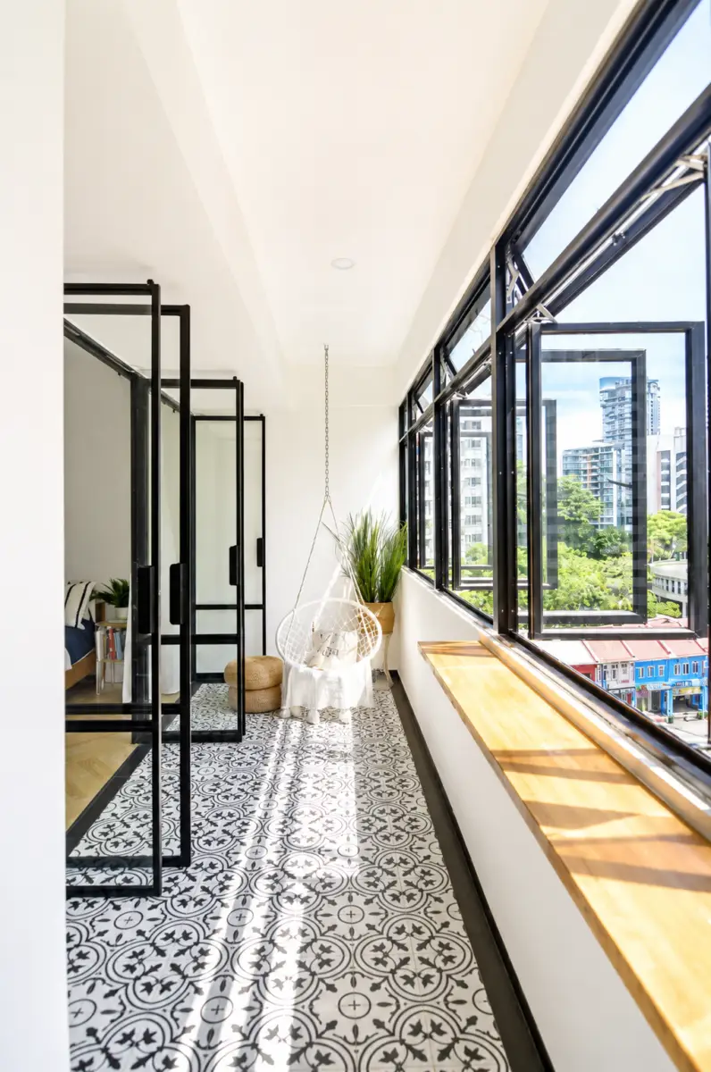 Sunlit balcony corner with a hanging wicker chair, patterned tile floor, and large windows overlooking the city.