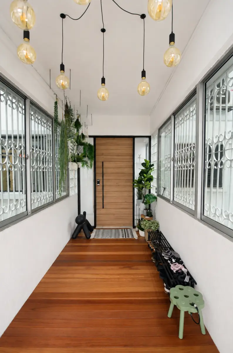 Narrow entry hallway with a warm wooden floor, hanging Edison bulbs, and potted plants along the left wall leading to a wooden front door. A shoe rack and small green stool sit on the right.