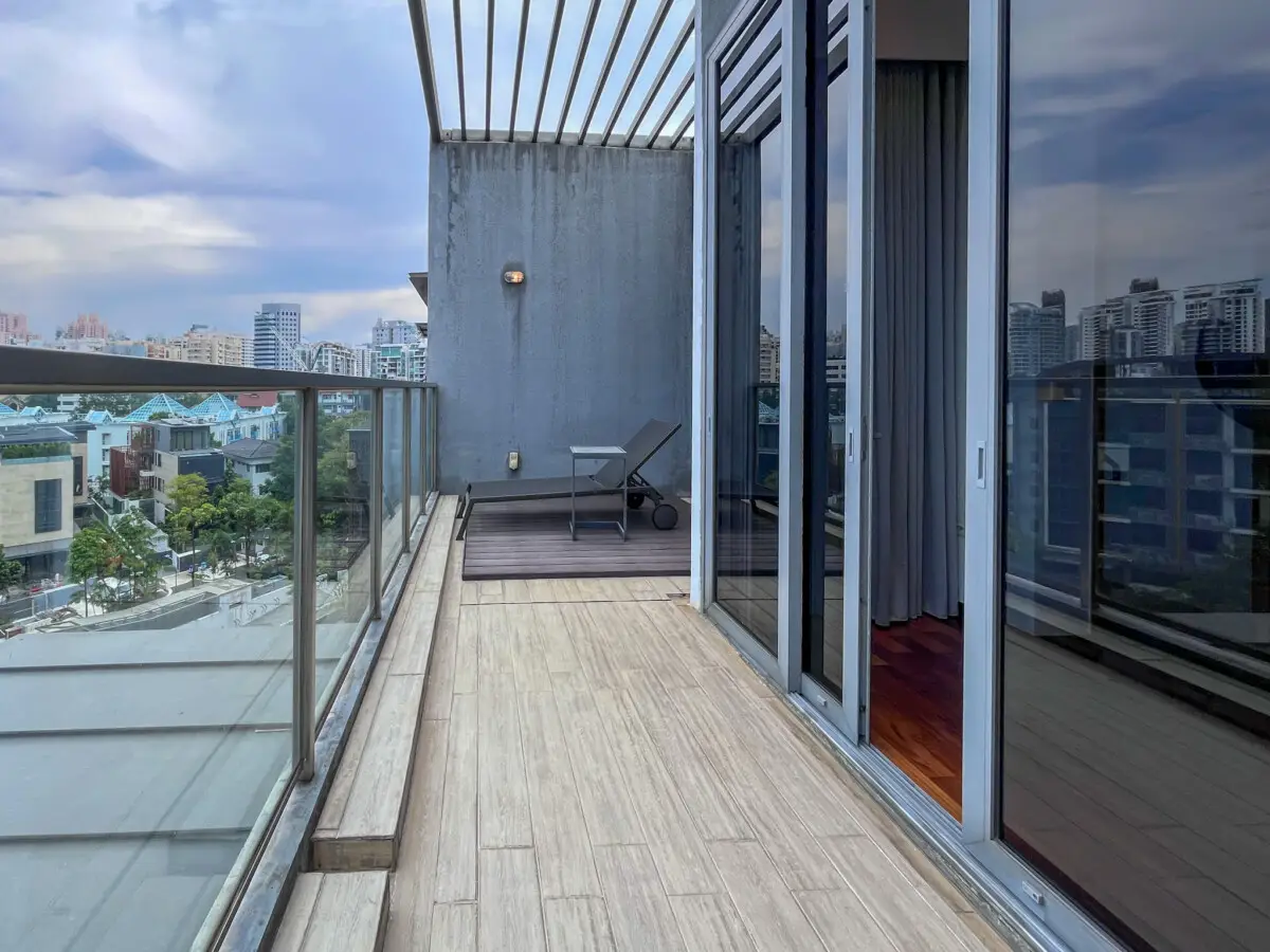Modern apartment balcony with glass railing, a lounge chair and small side table, overlooking a cityscape.