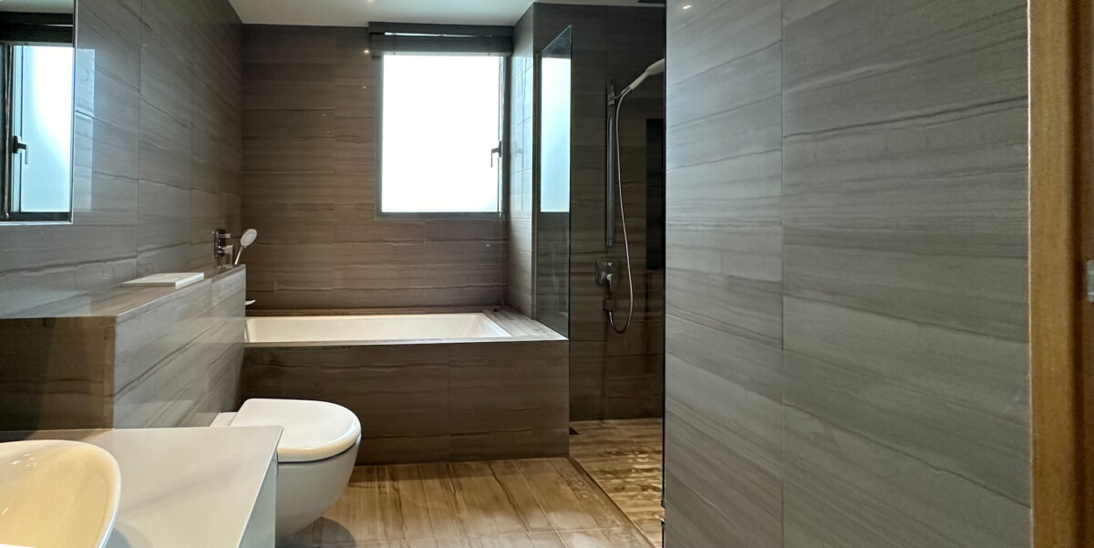 Modern bathroom with gray tiled walls, a built-in bathtub along the back wall, a wall-mounted toilet, and a glass-enclosed shower nearby.