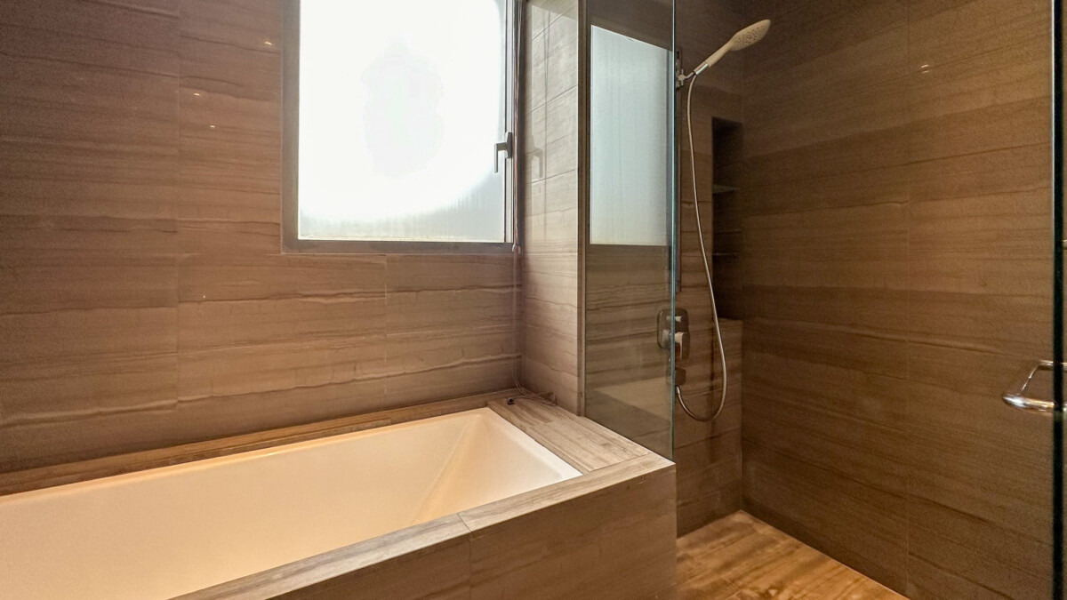 Modern bathroom with a white rectangular bathtub, wood-toned tiles, and a glass-walled shower area with a handheld showerhead.