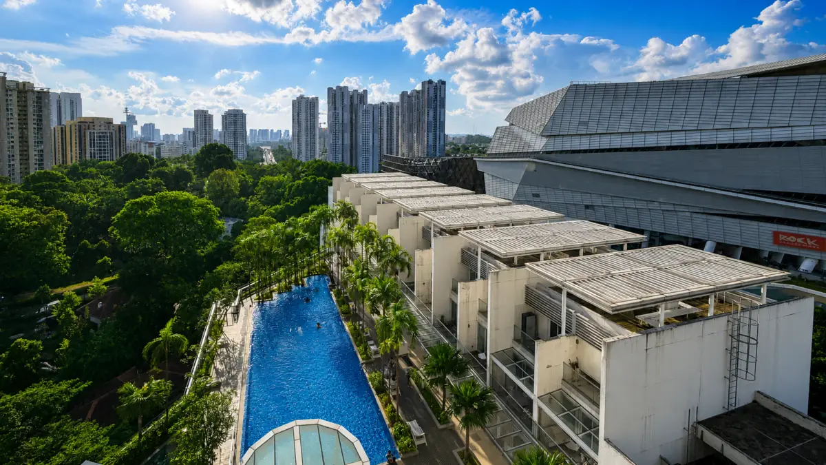 Rectangular outdoor pool with palm trees beside a white modern complex, city high-rises in the distance under a blue sky.