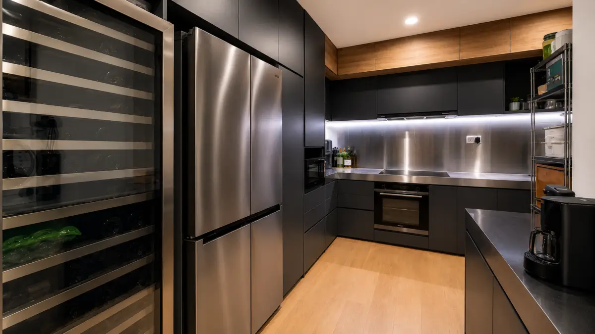 Modern kitchen with stainless steel refrigerator, wine cooler, black cabinets, and warm wood accents.