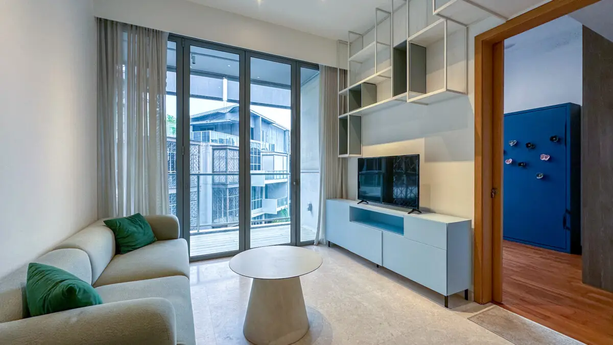 Bright modern living room with a beige sofa and green cushions, round white coffee table, and a TV cabinet beneath a geometric wall shelf beside glass doors to a balcony.