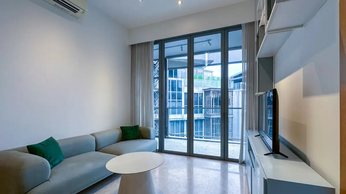 Modern living room with a light gray sofa and green cushions facing a TV on a white entertainment unit, large glass doors opening to a balcony, and a round white coffee table.