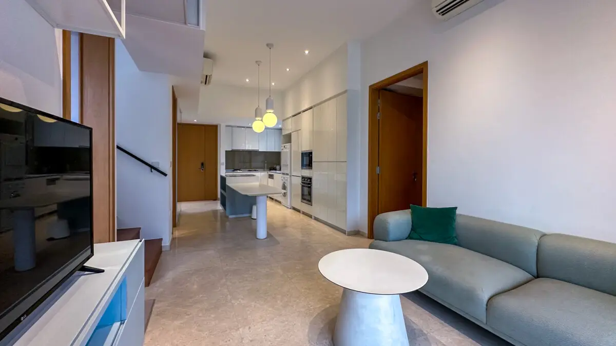 Open-plan living area with a gray sectional sofa, green cushion, round white pedestal table, and a bright white kitchen with an island in the background.