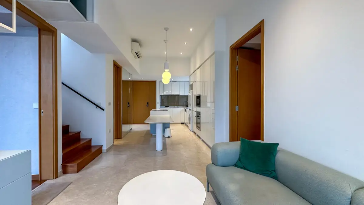 Open-concept living area with a gray sofa and green pillow, round coffee table, staircase on the left, and a white modern kitchen in the background.
