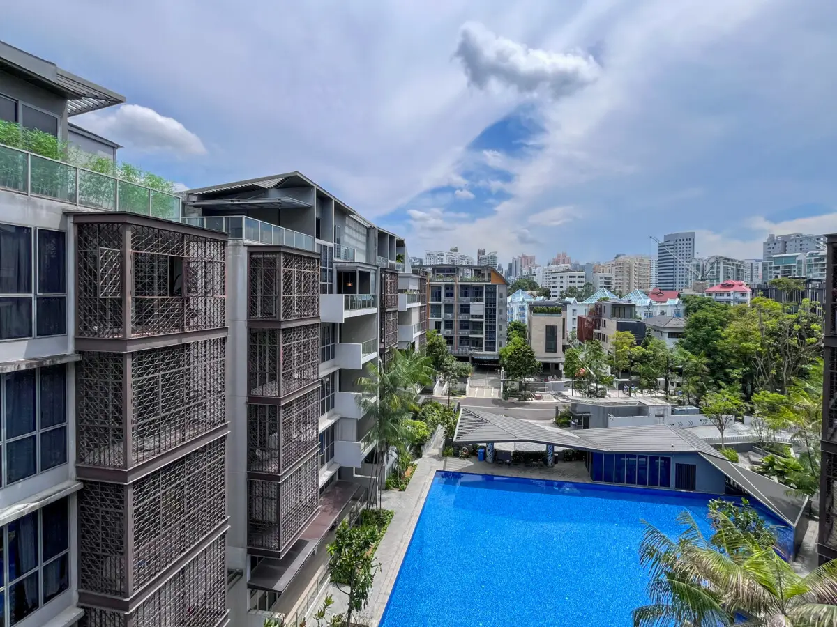 Aerial view of a modern apartment complex with a large blue swimming pool, latticed balconies, and a distant city skyline on a sunny day.