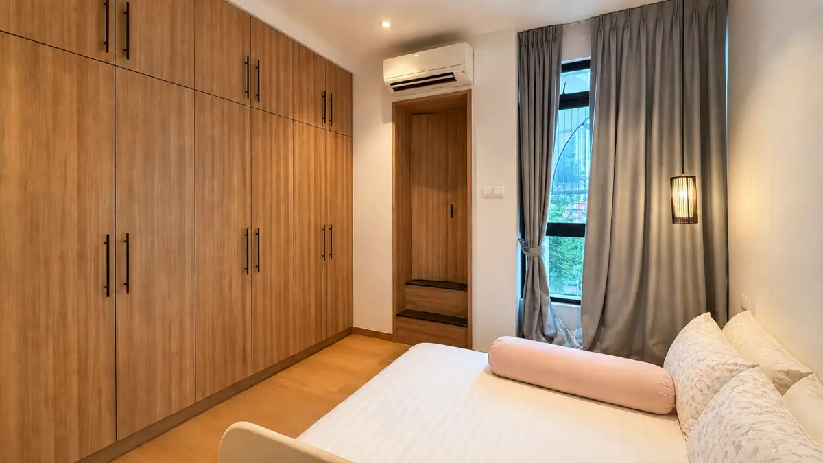 Bedroom with tall wooden wardrobes along one wall, a white bed with a pink bolster on the right, and gray curtains at a window.
