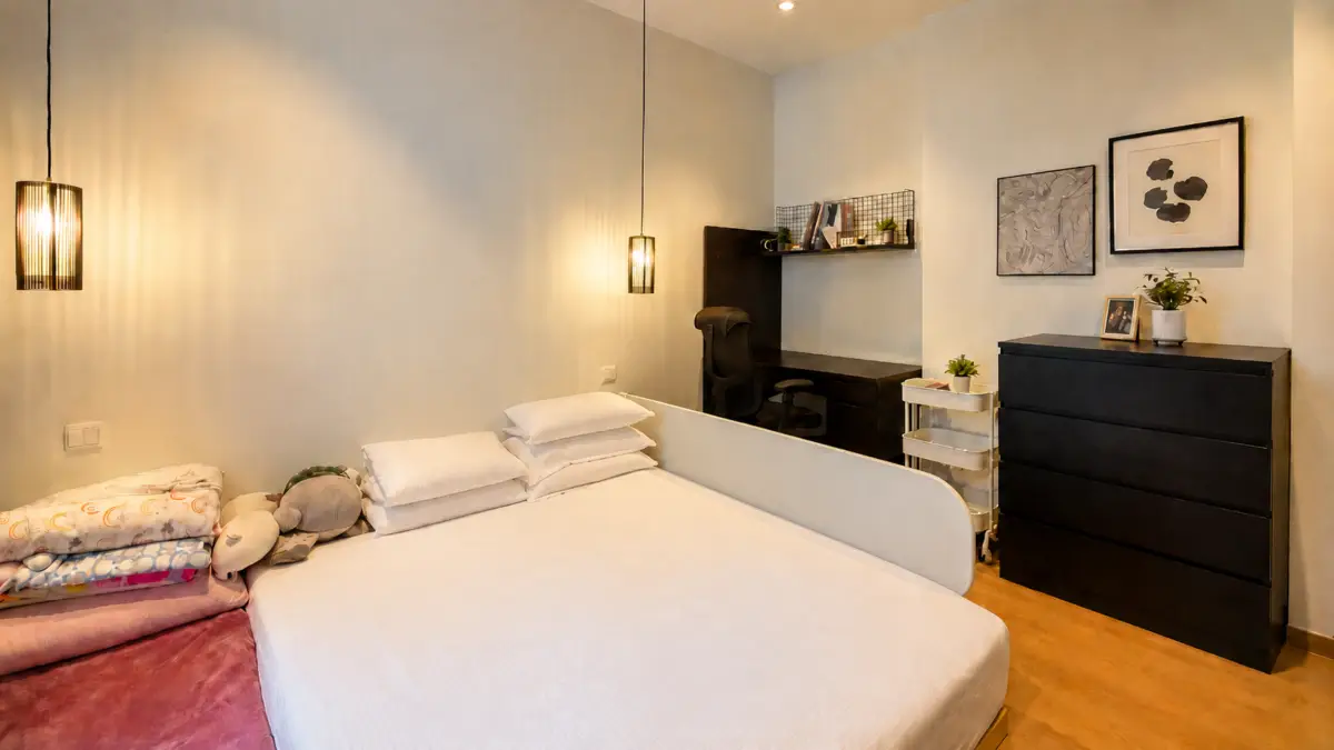Modern bedroom with a white bed, stacked pillows and plush toys, warm pendant lighting, and a dark dresser near a small desk area in the corner.