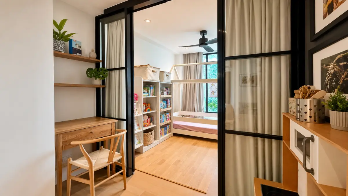 A bright children's study corner with a wooden desk and chair, open shelves, and a doorway to a playful bedroom with a bunk bed.