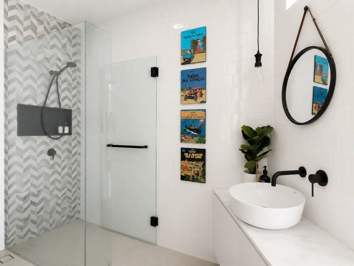 Modern bathroom with glass shower, white tile walls, a round mirror, and a white vessel sink on a marble counter.