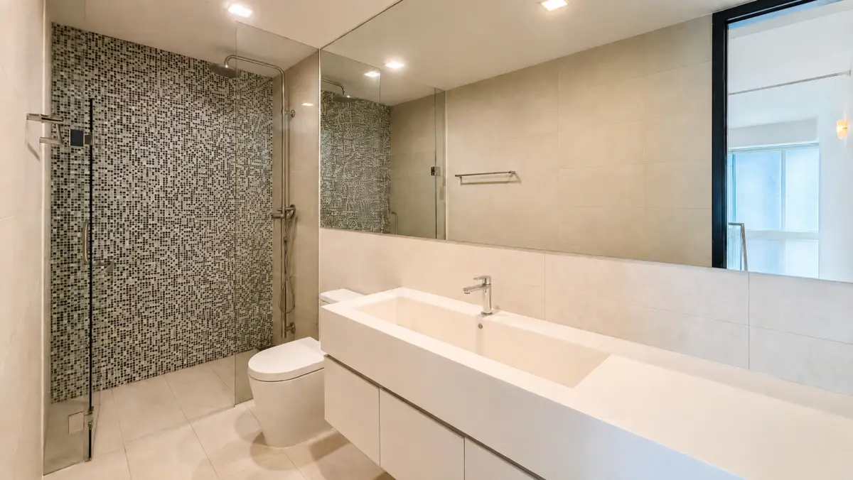 Modern bathroom with glass-enclosed mosaic shower, toilet, and a long white double-sink vanity with a large mirror above it.