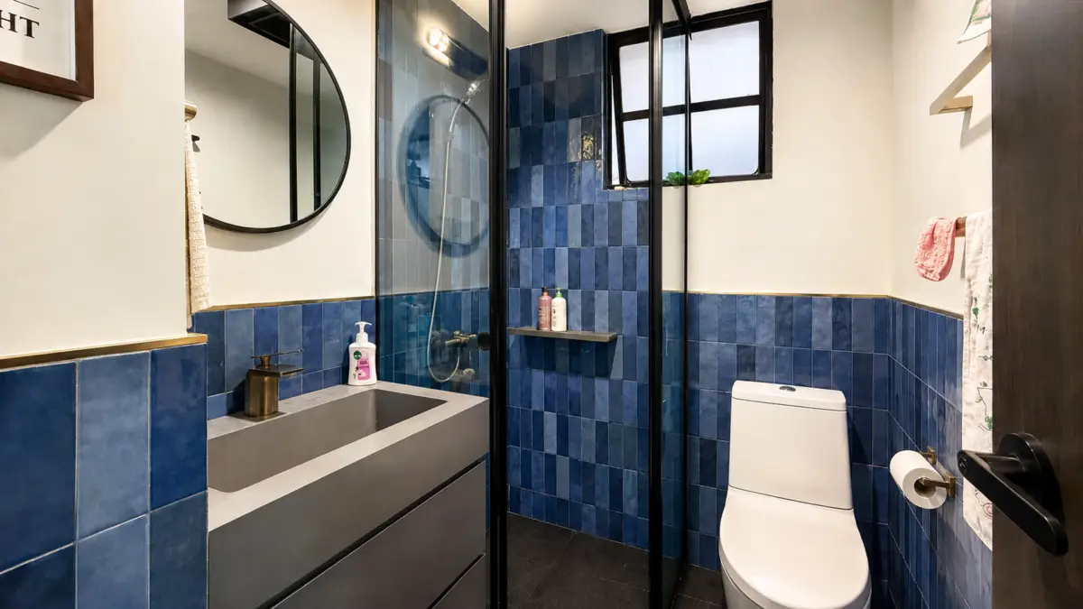 Modern bathroom with blue tiled walls, a gray vanity and round mirror beside a glass shower enclosure and white toilet.