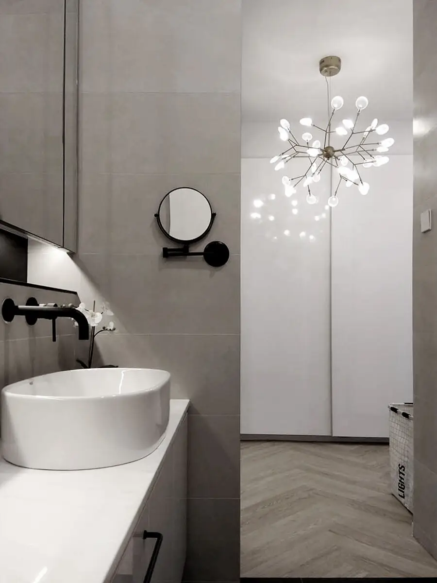 Modern bathroom with a round white vessel sink on a white countertop and black wall-mounted faucet, gray tiled walls, and a round vanity mirror. A decorative starburst chandelier hangs from the ceiling. Malming light and clean lines give a minimalist feel.