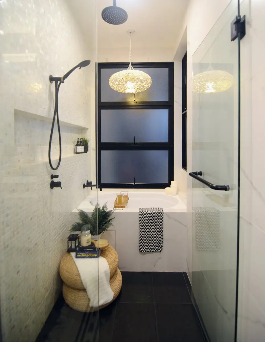 Modern bathroom with glass shower, black fixtures, and a frosted window; a wicker pouf with a white towel sits by the tub.